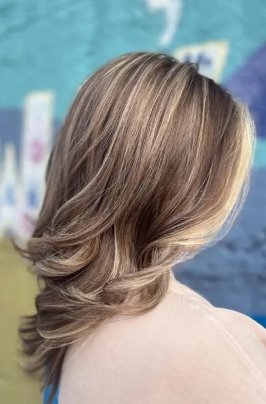Shoulder-length bronde hair with blonde ribbons through it, ends curled under, against a teal mural